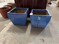 Two blue glazed square ceramic planters sitting on floor; one has green tape on the side and visible wear