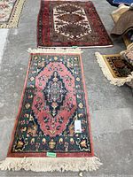 Photo showing two rugs laid on a concrete floor: front rug is floral pattern with pink and green colors, back rug is red with geometric design.