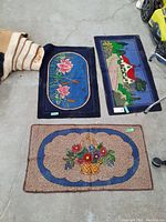 Photo showing three scatter rugs laid on concrete floor: water lily design, house and trees scenic design, and floral basket design.