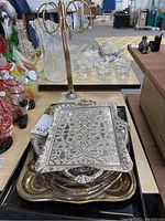 Multiple trays stacked: metal tray, silver-coated trays with floral designs, gold accented glass tray, on a table with decorative glassware in background