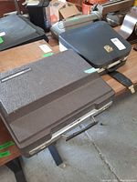 Two portable typewriters in closed carrying cases, one brownish-gray Smith Corona Coronomatic 2200 and one black Remington case with gold emblem, sitting on a table.