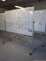 Full front view of large whiteboard filled with scheduling information written in multicolor marker, showing whiteboard surface, metal frame, and wheels.