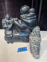 Photo showing both Inuit statues, black soapstone figurine and smaller chalkware figurine, positioned side by side on a flat surface with a dark background.
