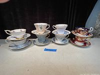 Full set of 8 tea cups and saucers stacked and arranged in four groups, showing the variety in color and pattern. Mainly white with floral and ornate gold designs, plus richly colored cobalt blue and red pieces.
