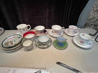 Overview of the lot showing multiple cups and saucers in different styles and patterns arranged on a table.