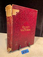 Photo of the front cover of an antique book titled 'Queen Victoria' showing red embossed design and damaged spine.