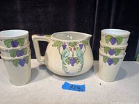 Full view of antique Limoges pitcher and six matching juice glasses with bird and grape cluster design placed on a surface.
