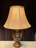 Full view of gilded table lamp with beige silk fabric shade on a white surface against black background.