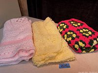 Three folded handmade crocheted blankets in pink, yellow, and multicolored floral pattern colors shown on a dark background.