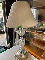 Full view of the large vintage crystal table lamp with a beige pleated fabric shade on a metal base with the crystal body and hanging prisms visible.