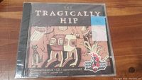 Front view of the sealed jewel case for The Tragically Hip CFL Grey Cup commemorative CD. Shows cover art with abstract figures and CFL Grey Cup logos.