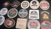 Photo showing 14 different vintage cardboard beer coasters with various brewery logos and designs arranged flat for display.