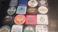 Photo of 16 vintage cardboard beer coasters with various brewery logos and designs on a dark surface.