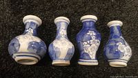 Four vintage ceramic bud vases arranged side by side on a dark carpet. The vases are white with blue floral designs, vary slightly in shape but consistent in size and decoration style.