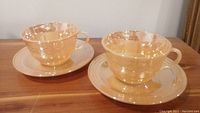 Two pairs of translucent peach colored Fire King tea cups each paired with matching saucers on wooden surface.