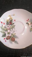 Close-up of saucer showing floral winter pattern with pine cones, holly leaves, red berries, and scalloped gold trim.