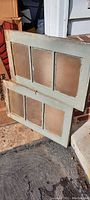 Photo showing two antique basement windows painted gray with three vertical glass panes each, leaning against a wall outdoors.