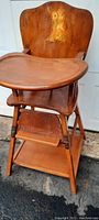 Full view of the antique highchair showing carved wooden backrest with painted figure, attached tray, footrest, and lower shelf. The finish shows light age-related wear.
