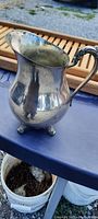 Side view of the antique silver-plated three-legged pitcher showing tarnish, curved spout, and ornate handle.