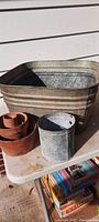 Front view of a large rectangular galvanized tub, a small galvanized bucket, and five terracotta flowerpots stacked on a white table.