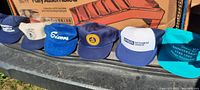 Seventeen vintage advertising trucker hats displayed outdoors, showing various logos and styles, including blue, green, white, red, and teal colored hats with business advertisements.