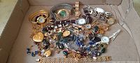 Box containing assorted costume jewelry items including pins, necklaces, earrings, pendants, bracelets, and watches, mixed and overlapping.