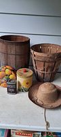 Photo showing entire lot of two wooden peck baskets, a large wooden nail keg, three metal tins including Hershey's Cocoa, and a straw hat.