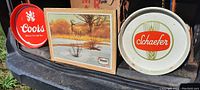 Lot showing round metal trays with Coors and Schaefer logos flanking a framed cardboard sign depicting a deer scene.
