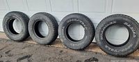 Four Goodyear Wrangler tires standing side-by-side with raised white lettering visible on outer sidewalls and black wall on the other side.