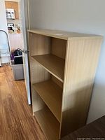 Front and side angle view of a light wood-tone pressboard bookcase showing three shelves and peg holes for adjustable shelves.