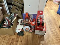 Wide photo showing various Coca-Cola collectables including cookie jar, fridge, tins, signage, ornaments on wood floor