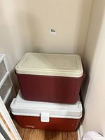 Two coolers stacked, smaller solid red cooler with beige lid on top of large Rubbermaid cooler with white lid and latch.