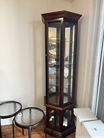 Front side view of tall glass curio cabinet with wooden frame placed near a wall and two glass top side tables next to it.