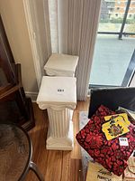 Two white matching decorative pillars shown standing side-by-side on hardwood floor near window and other household items.