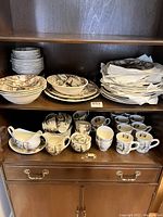 Photo shows shelves holding a variety of ceramic dishes including plates stacked together, bowls, cups, a gravy boat, and other small dishes. All pieces match showing a scenic vintage pattern.