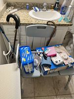 Adjustable shower chair with gray padded seat and backrest, various small medical items on seat including a box of adult incontinence products, a blood pressure cuff, and other medical supplies. The chair has metal frame and armrests with black handles.