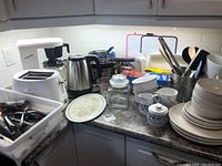 Wide view of corner kitchen countertop with T-Fal kettle, Sunbeam toaster, black coffee maker, assorted pots, dishes, glass jar, canisters, and stack of white and blue patterned plates and bowls