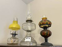 Three oil lamps shown together, small clear glass with yellow top, large clear glass lamp, decorative stained glass style lamp