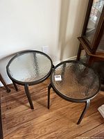 Two matching outdoor side tables with metal frames and textured glass tops, shown on a wooden floor indoors.