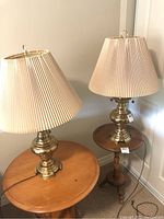 Photo showing the pair of brass table lamps with beige pleated shades on wooden pedestal tables, displaying both lamps and their proportions.