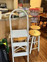 White metal step ladder with three textured steps and curved top handle, two wooden stools with natural wood seats and white painted legs behind ladder, kitchen background.