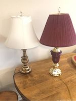 Pair of table lamps on wooden table; left lamp has brass base and off-white bell shade, right lamp has brass-look/maroon base with maroon pleated shade