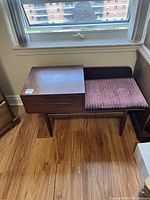 Full front view of wooden telephone table with drawer and mauve fabric cushioned seat against a window background.