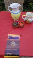 Two glass jugs and a fruit basket cream and sugar holder displayed on a red cloth with a Rotary Club banner in front, outdoor setting