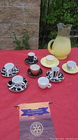 Photo of five assorted espresso cup and saucer sets in colorful designs, three extra saucers, and a yellow glass pitcher on a red tablecloth outdoors.