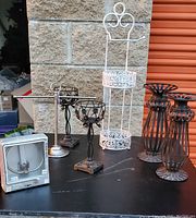 Photo of multiple decorative candleholders in black and bronze tones, a white decorative two-tier toilet paper holder, and several bronze-toned toilet paper holders on a table against a stone and orange background.