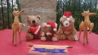 Five holiday figurines including three bears and two reindeer positioned on red fabric with an outdoor stone-wall background.