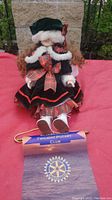 Front view of Christmas doll sitting on a red surface with part of Rotary Club plaque visible.