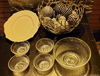 Photo of large decorative metal bowl with multiple black and white patterned decorative balls, four small clear ribbed glass bowls, one large clear ribbed glass bowl, and one white glass platter on a dark reflective surface.