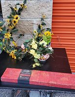 Photo showing a rectangular magnetic wall board with red and black pattern, a faux flower arrangement in a decorative bowl, and a long yellow flower vine against a stone wall and red door background.